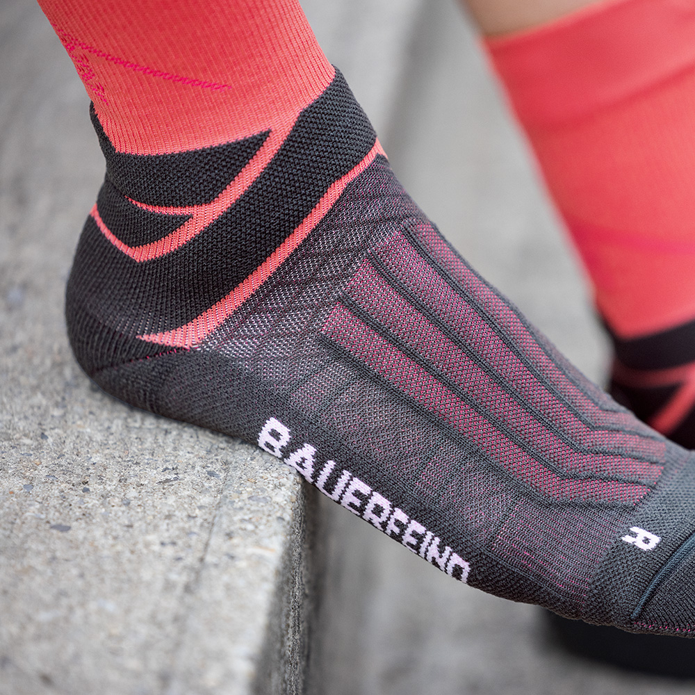 Close-up of a black running sock with red accents by Bauerfeind, worn while sitting on a concrete step. The sock displays the integrated compression pattern and the Bauerfeind lettering on the side.