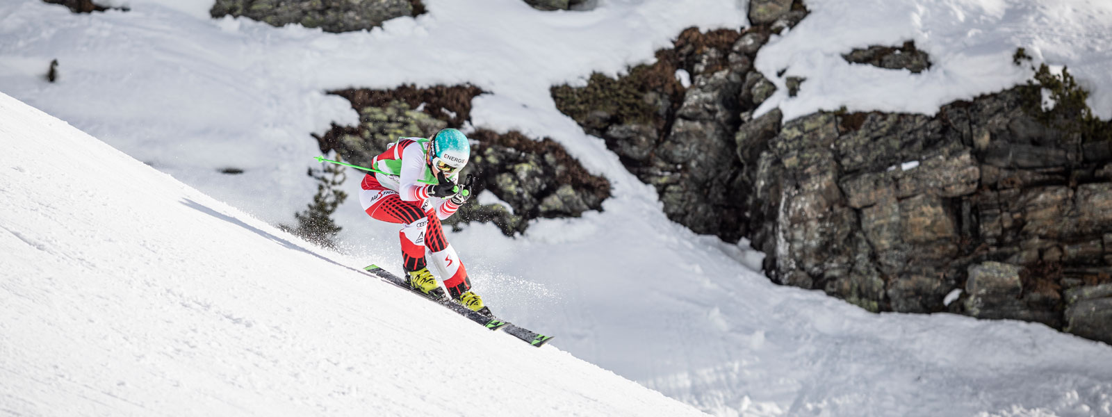 Off to the downhill: enjoy your winter on the slopes and with our products Ski runner in a red and white racing suit in a downhill position with a slight swing with large snow-covered rocks in the background