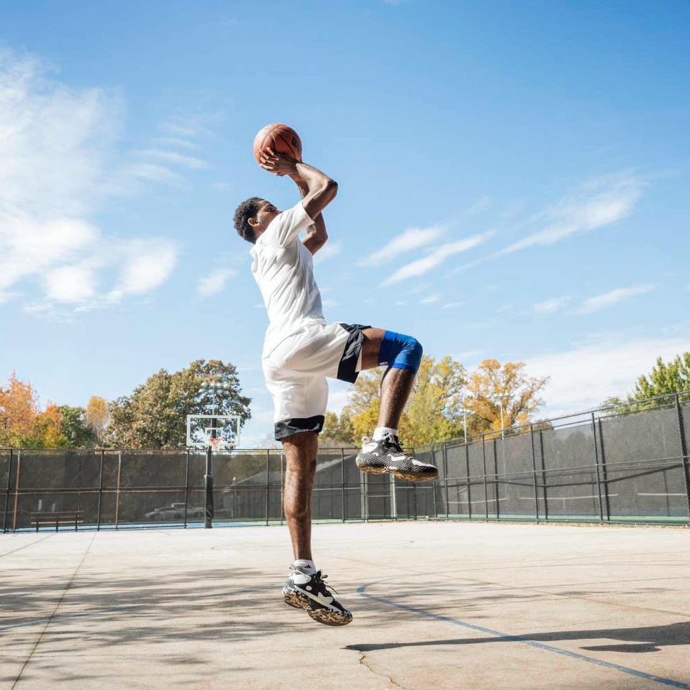 A basketball player wearing a blue Dallas Mavericks knee support while taking a jump shot on an outdoor court. The brace supports the knee during movement.