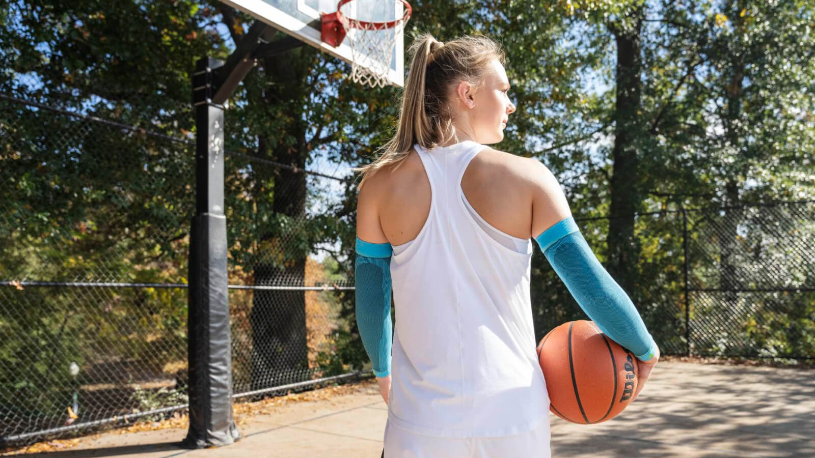 A female basketball player stands on an outdoor court, holding a basketball in her right hand. She is wearing light blue compression arm sleeves and looking focused toward the hoop.