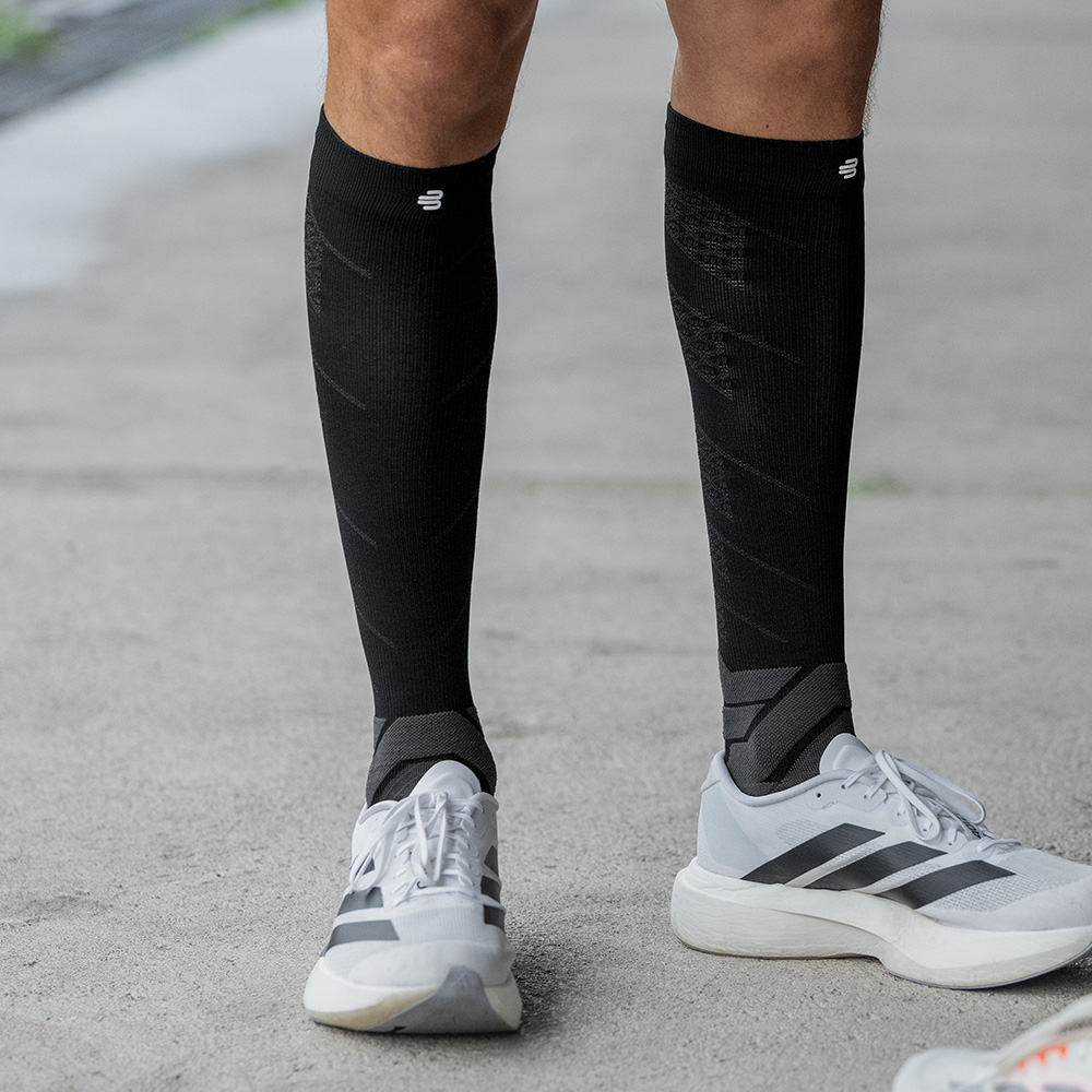 A sporty man stands facing forward on asphalt, wearing black Bauerfeind compression socks and white running shoes. His upright posture and visible calf tension emphasize the socks’ stabilizing effect during standing.