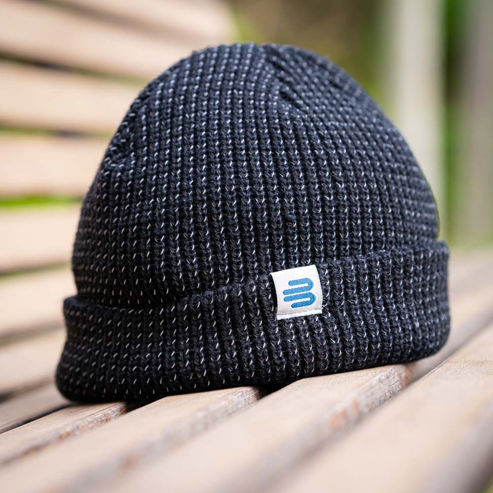 A black Bauerfeind Sports knitted beanie with the logo, lying on a wooden bench outdoors. The chunky knit gives it a modern, sporty look.