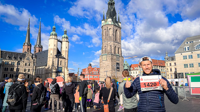 A participant of the Mitteldeutscher Halbmarathon holds up his bib number on Halle's (Saale) marketplace. Behind him, the historic Roter Turm and Marktkirche are visible. In the foreground, runners and spectators prepare for the race.