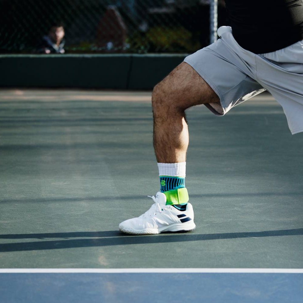 A tennis player on the hard court, moving while serving. He wears the Bauerfeind Sports Ankle Support Rivera in blue-green over his white sports socks.