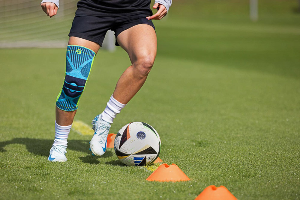 A football player training on the field, wearing a Bauerfeind knee brace. The focus is on the stability and support provided by the brace during dynamic movements, such as dribbling around cones.