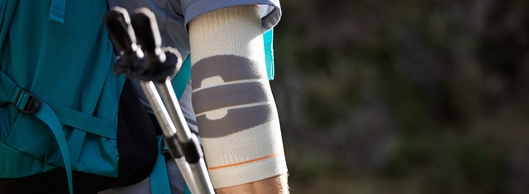 A hiker wearing a backpack uses trekking poles on a narrow outdoor trail. An Outdoor Compression Elbow Support is worn on the elbow, providing secure support and stability during long hikes and uneven terrain.