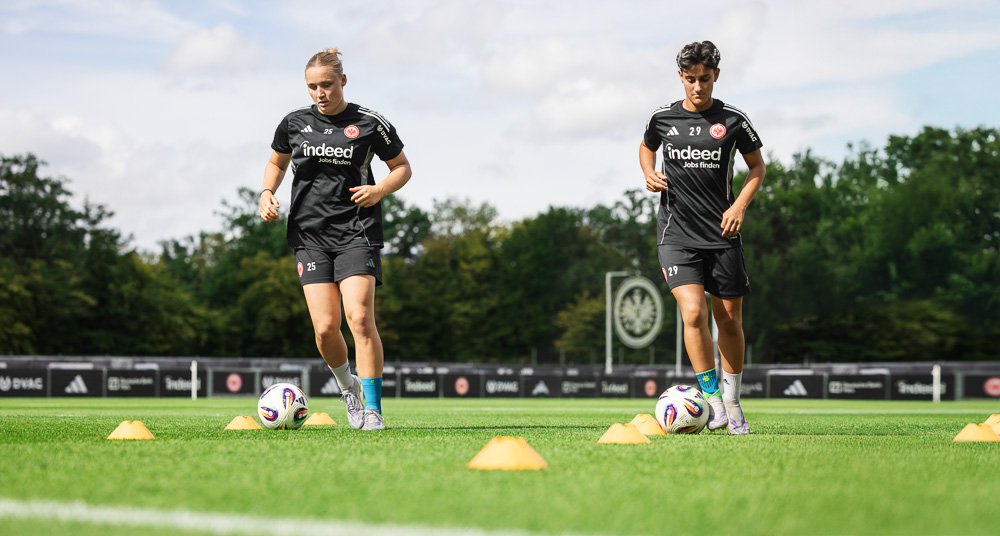 Two Eintracht Frankfurt players are training on the pitch with cones and a football, wearing black athletic outfits. Both are equipped with Bauerfeind Sports compression gear, suited for dynamic movements.