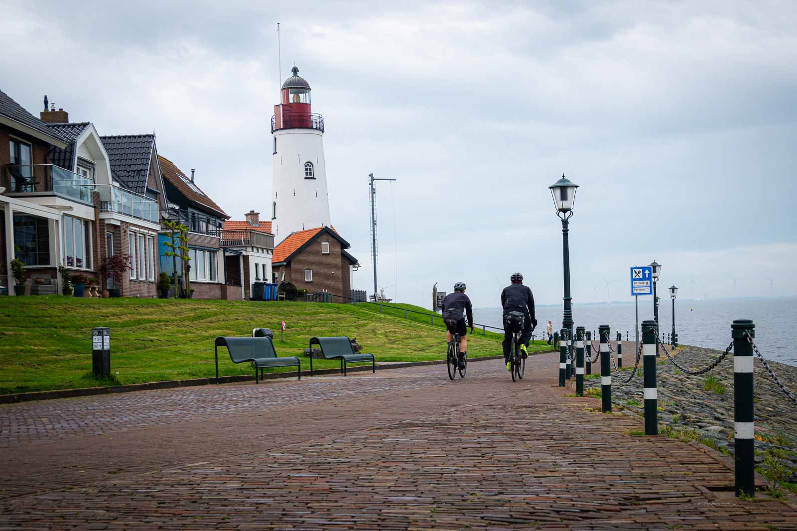 Two ultra-cycling cyclists from behind riding along a promenade where a lighthouse stands.