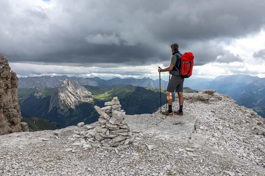 A person with a red backpack stands on a rocky ledge in the Dolomites, gazing into the distance. The sky is cloudy, and there is a cairn structure in the foreground.