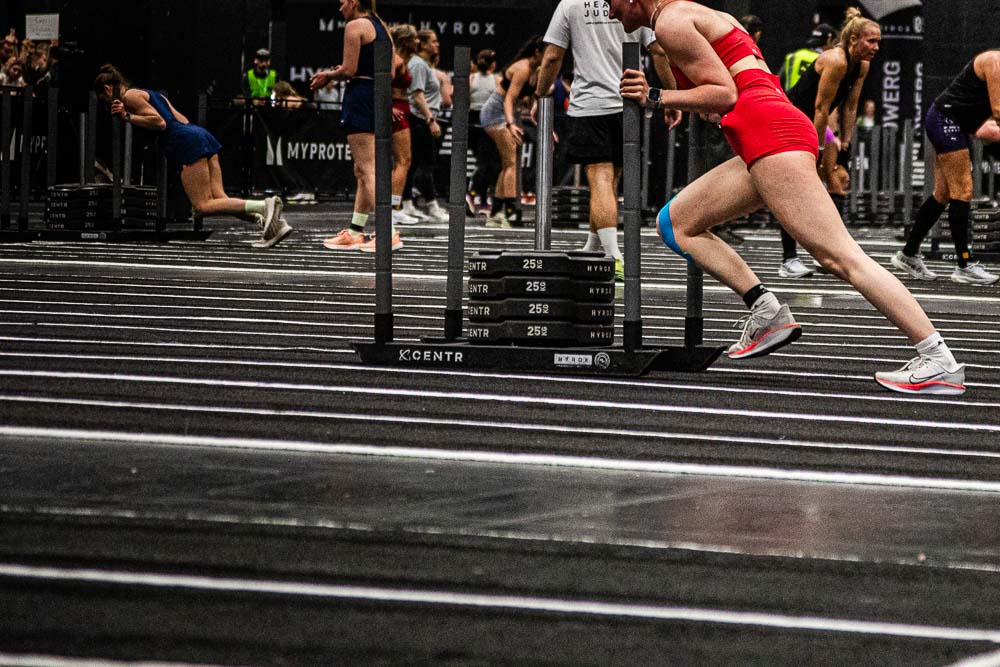 Athlete in a red sports outfit pushing a heavy sled across the marked indoor workout area during a HYROX competition, while other participants work at their stations in the background; the blue kinesiology tape on the thigh