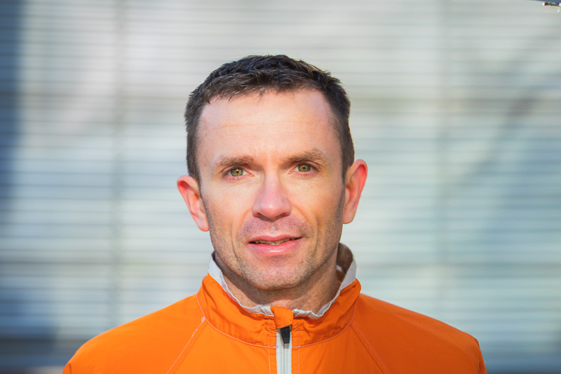 A man in a bright orange jacket smiles at the camera. The background features a modern building with trees and blooming plants.