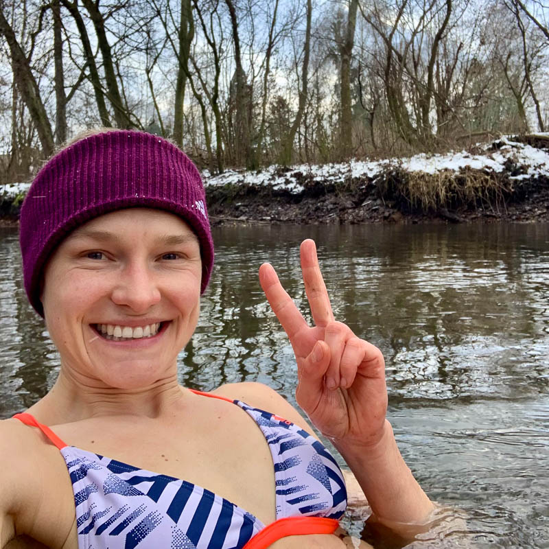 Anna Hahner is standing chest-deep in cold water, surrounded by a snowy landscape and bare trees, smiling and showing a peace sign with her fingers.