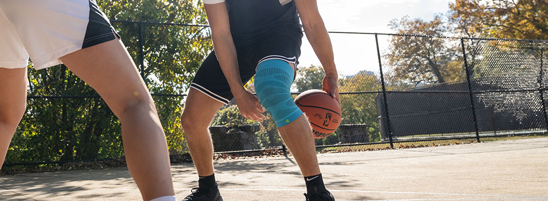 A basketball player stands on a concreted court that is surrounded by a fence and performs a trick with the ball by passing it through his two legs. Another player is standing opposite him. He is wearing a knee sleeve.