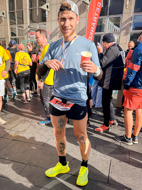 Standing proudly at the finish line after completing the half marathon in Halle, with a medal and a drink in hand, savoring the moment post-race.