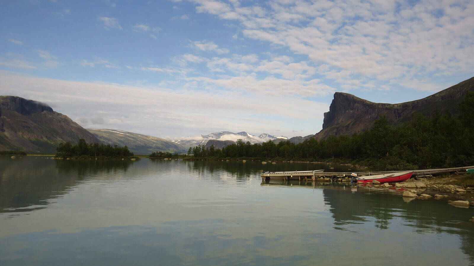 Panorama of a lake surrounded by trees and small mountains on the right edge is a small boat on the jetty