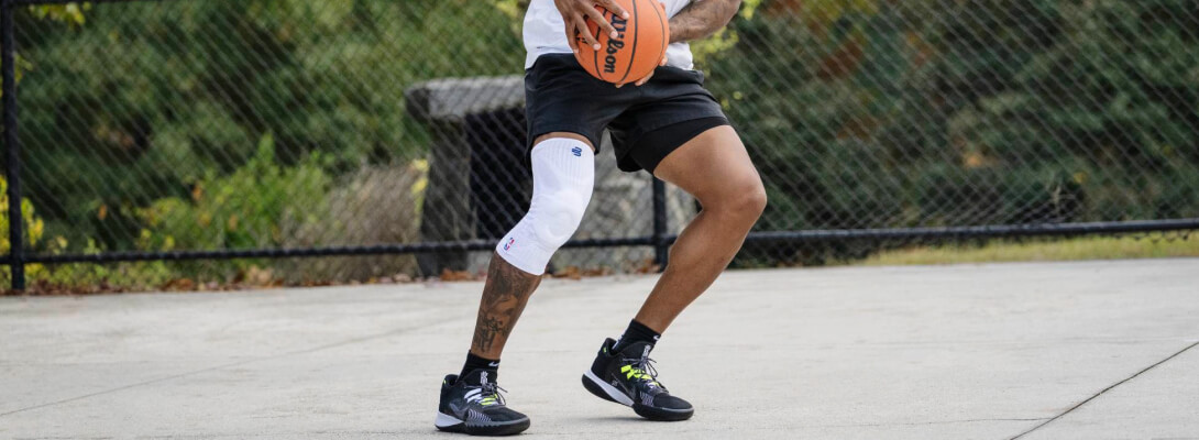Basketball player wearing a white compression knee support while handling the ball on an outdoor court. The white brace contrasts with his skin tone and black shorts, displaying a visible NBA logo.