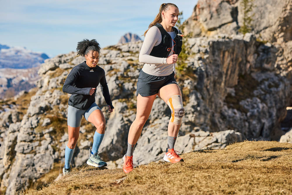 Two female athletes run uphill on a narrow mountain trail. The lead runner wears a light-colored outdoor compression knee support, while the second athlete follows closely. Rocky terrain and an alpine landscape frame the scene.