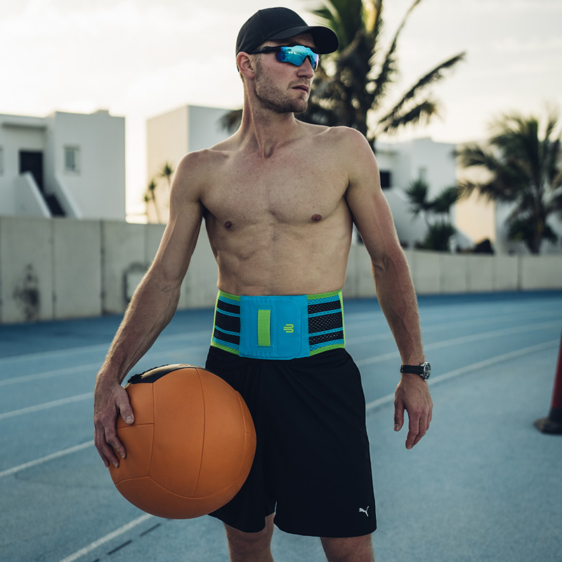 A sporty man wearing a baseball cap and sunglasses stands on a running track wearing a blue back support with green accents. He holds a large medicine ball in his hand.
