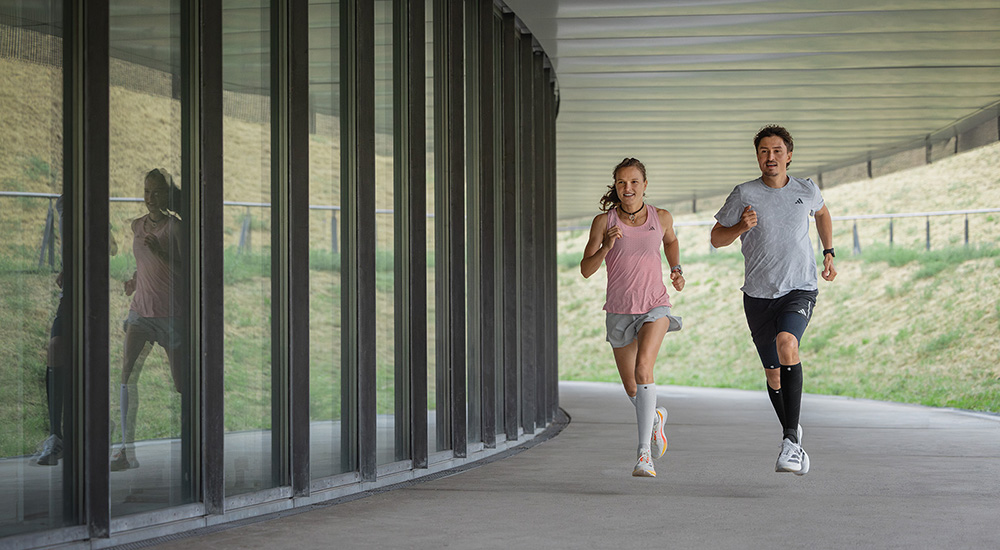 Two runners – a woman and a man – are running together under a covered passage. The woman wears white compression sleeves, the man black ones. Both move in sync, looking focused and full of energy.